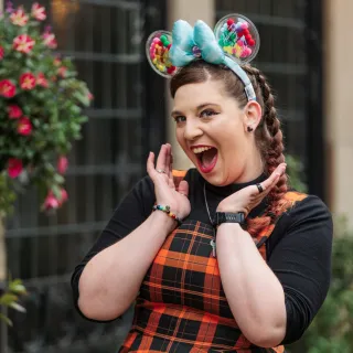 Rachel looks excitedly towards the camera, hands framing her face whilst wearing a black and orange tartan dress with a black long sleeved under top, and a pair of 'UP' themed Mickey ears