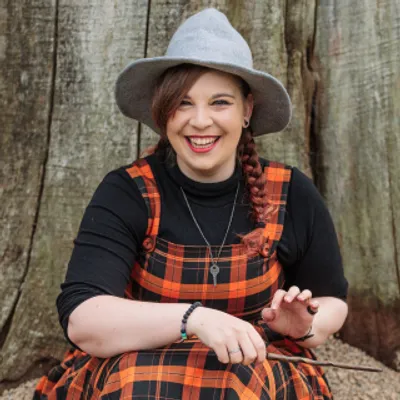 Rachel is sat in front of a large tree with a big smile on her face, wearing an orange tartan dress, grey witches hat, TARDIS key necklace and holding a wand