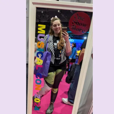 Mirror selfie of Rachel smiling wearing Wicked themed leggings, a Beetlejuice style jacket, a 'The Show Must Go On' top, and doc martins with rainbow laces. The mirror has 'One Show More' and 'Musical Con 2024' on it.