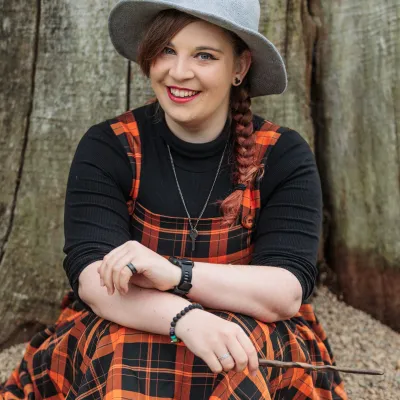 Rachel smiles to the camera whilst wearing a black and orange tartan dress with a black long sleeved under top, a grey witches hat and holding a wand