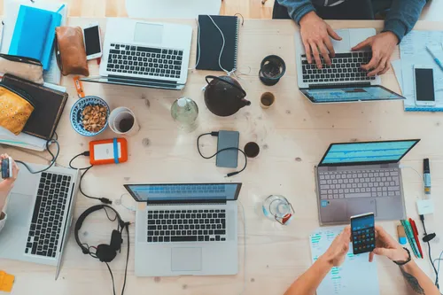 worktop with two people working at multiple laptops with cups of coffee, cables, phones and headphones