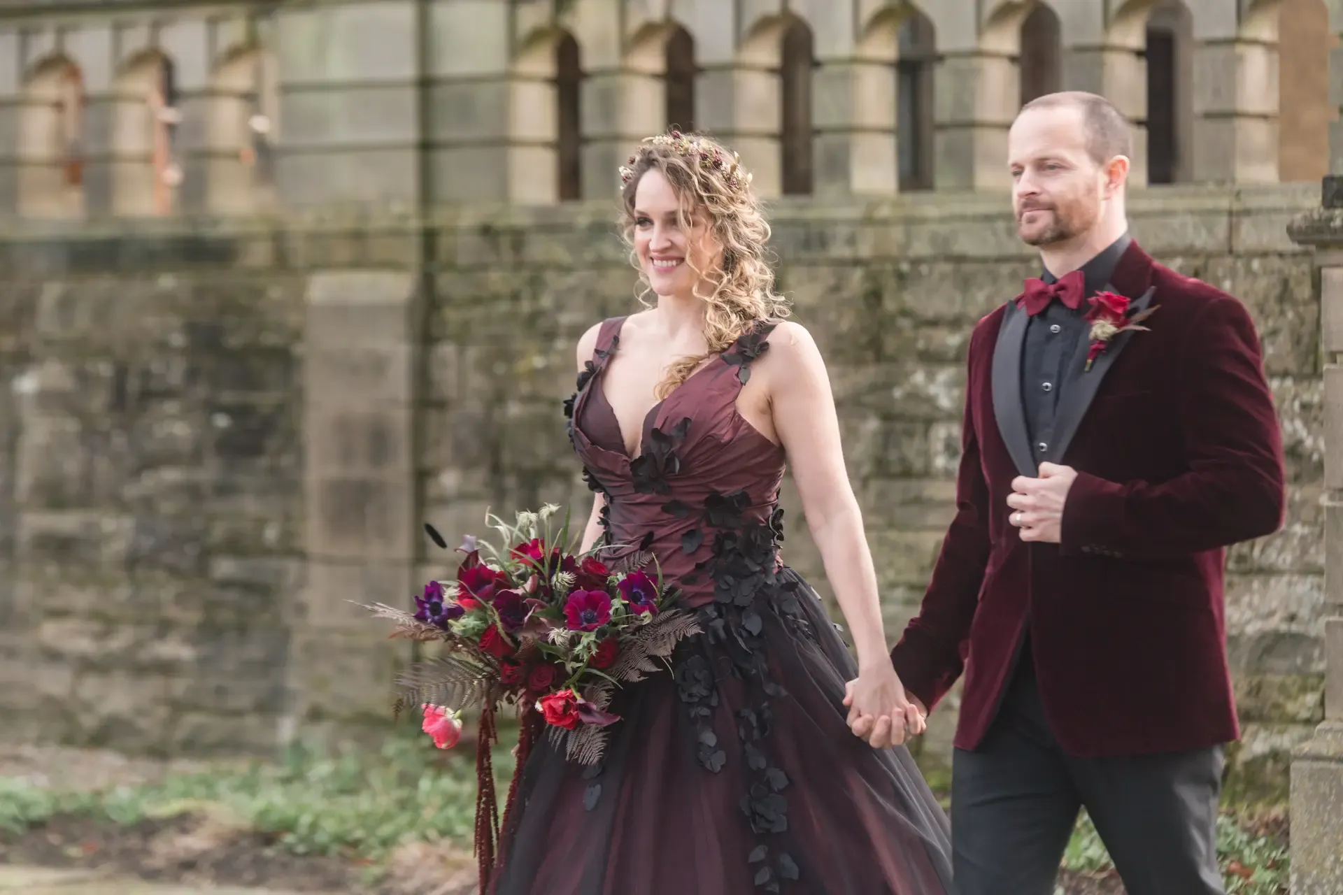 Bride and groom walking in the courtyard of Fanhams Hall. The groom is wearing a red velvet jacket, red bow tie, a black shirt and trousers with a red flower buttonhole. The bride is wearing a bespoke dark red wedding dress with black flower embellishments and black netting overlay, her bouquet is made up of deep red and purple florals with red fern and other foliage.